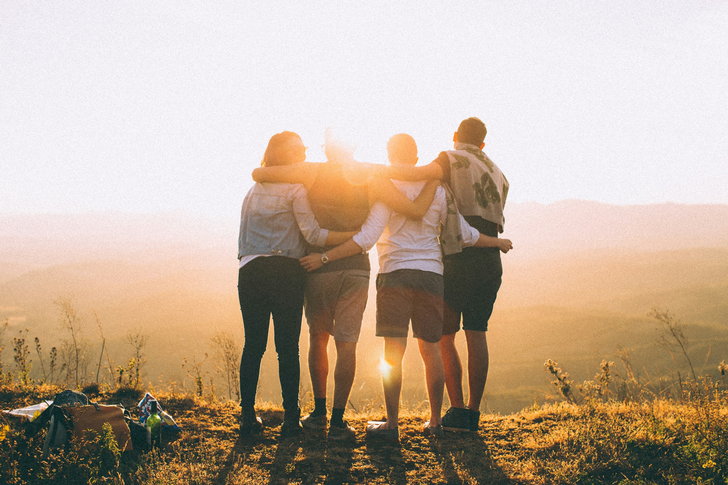 Four young adults stand on a hill watching the sun rise with their backs to the camera and their arms around one another.